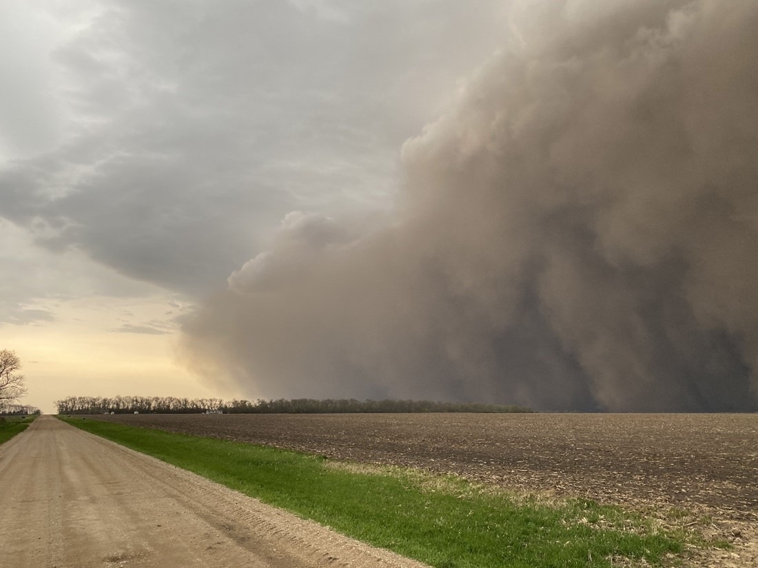 A deep, black storm cloud--a derecho--rolls over the righthand side of the landscape, lifting dirt and debris from the farm field covering the image to the horizon. 