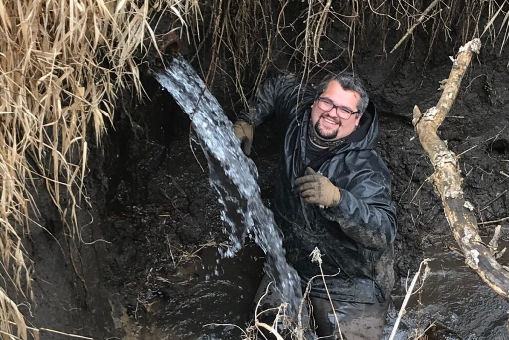 A smiling man looks up from a muddy pit as water flows from a tile drain above him.