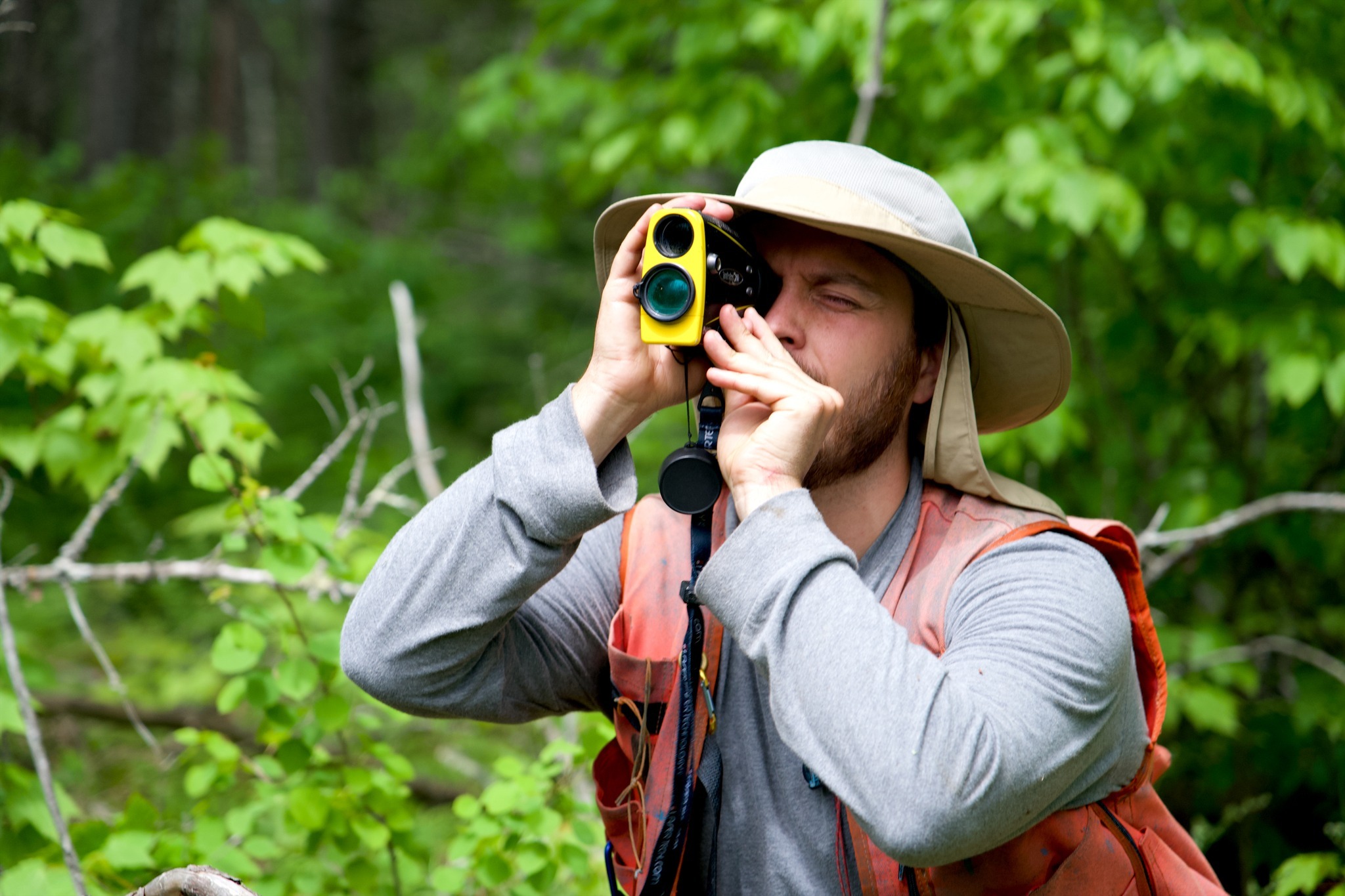 A man in an orange vest stands in the forest, sighting trees through a small, scope-like object. 